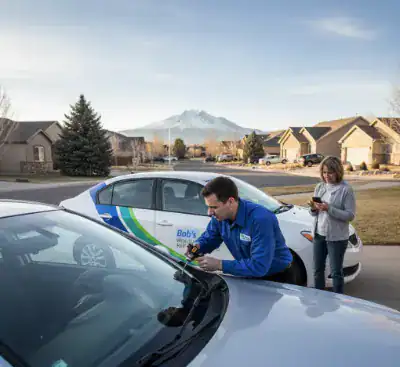 Mobile windshield technician repairing a chip at a Colorado Springs home while customer reviews insurance options on a phone.